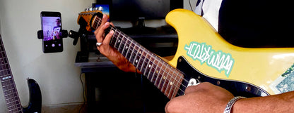 First-person POV of a musician playing an electric guitar with a headstock-mounted phone holder recording a video of the fretboard performance.